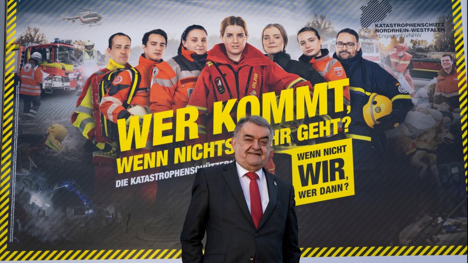 Minister Reul vor dem Plakat "Wer kommt, wenn nichts mehr geht" 