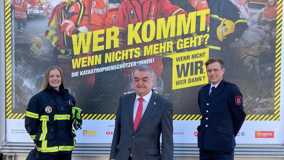 Minister Herbert Reul mit zwei Ehrenamtlichen vor dem Plakat "Wer kommt, wenn nichts mehr geht" 