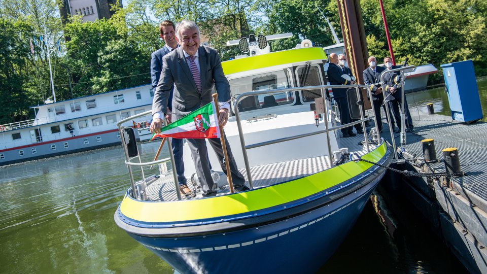 Herbert Reul lachend vorne auf dem neuen Boot der Wasserschutzpolizei mit NRW-Flagge in der Hand