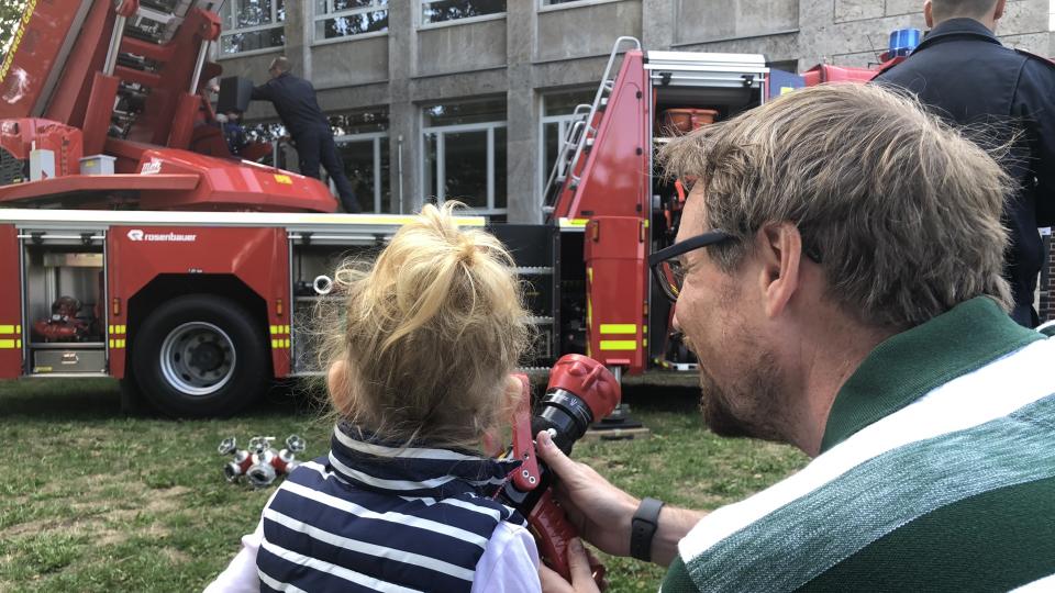 Ein kleines Mädchen schaut mit ihrem Vater auf einen Feuerwehrwagen und hält einen Feuerwehrschlauch