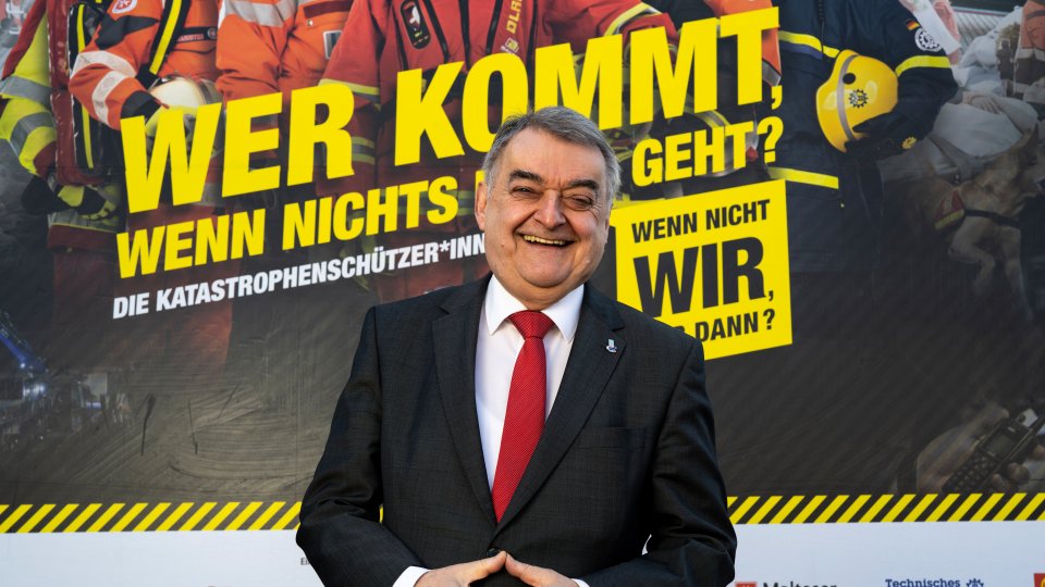 Minister Herbert Reul lachend vor dem Plakat "Wer kommt, wenn nichts mehr geht" 