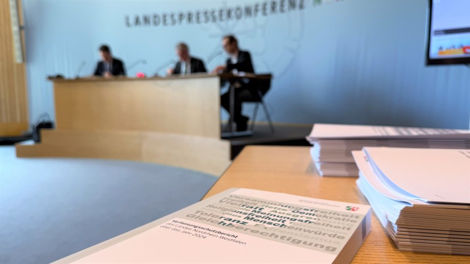 Pressekonferenz, im Vordergrund der aktuelle Bericht, im Hintergrund drei Herren