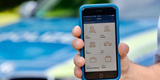 Eine Hand hält Smartphone in die Kamera, im Hintergrund ein unscharfes Polizeiauto